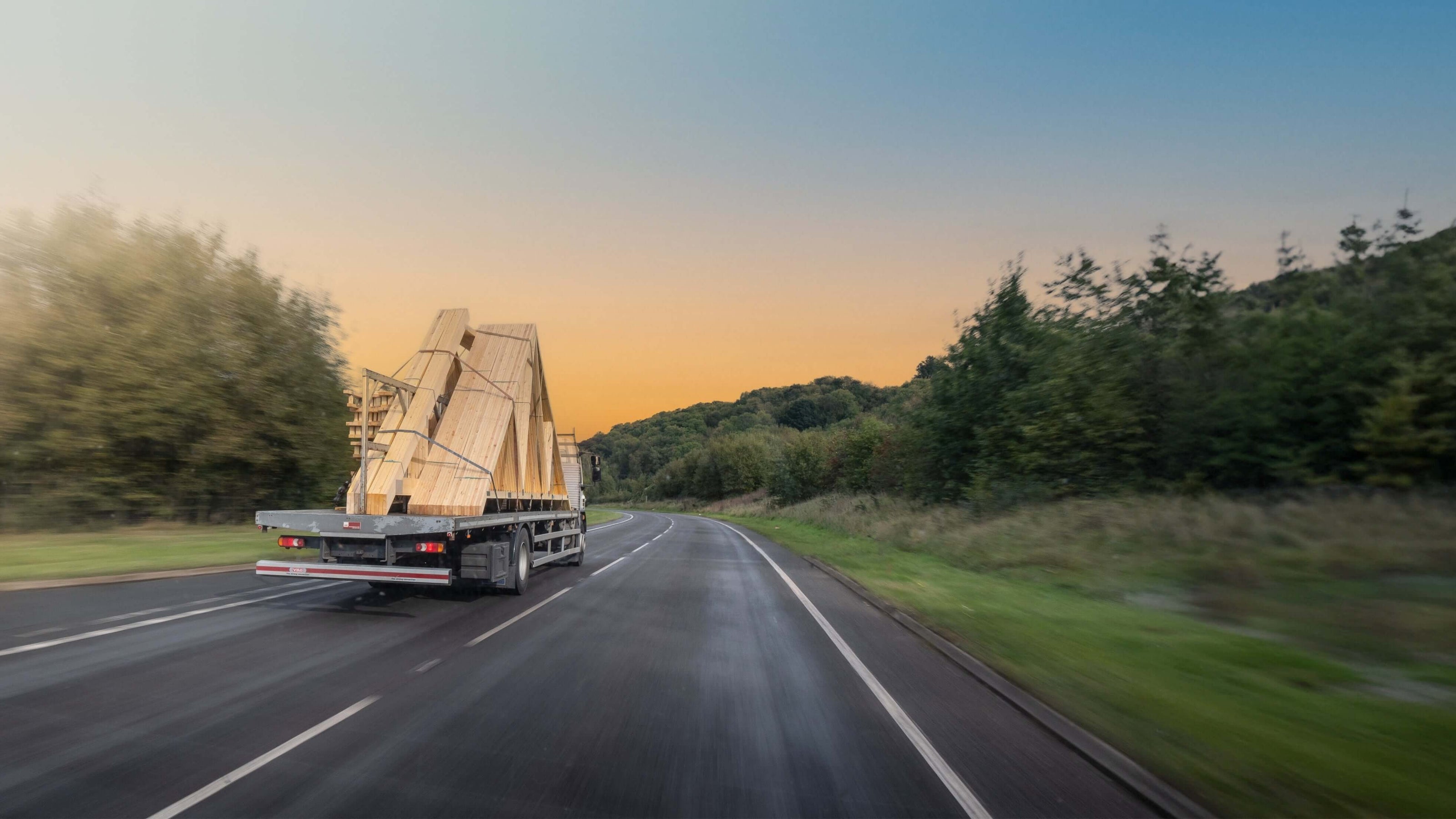 Truck transporting large wooden trusses on a road with trees and sunset in the background