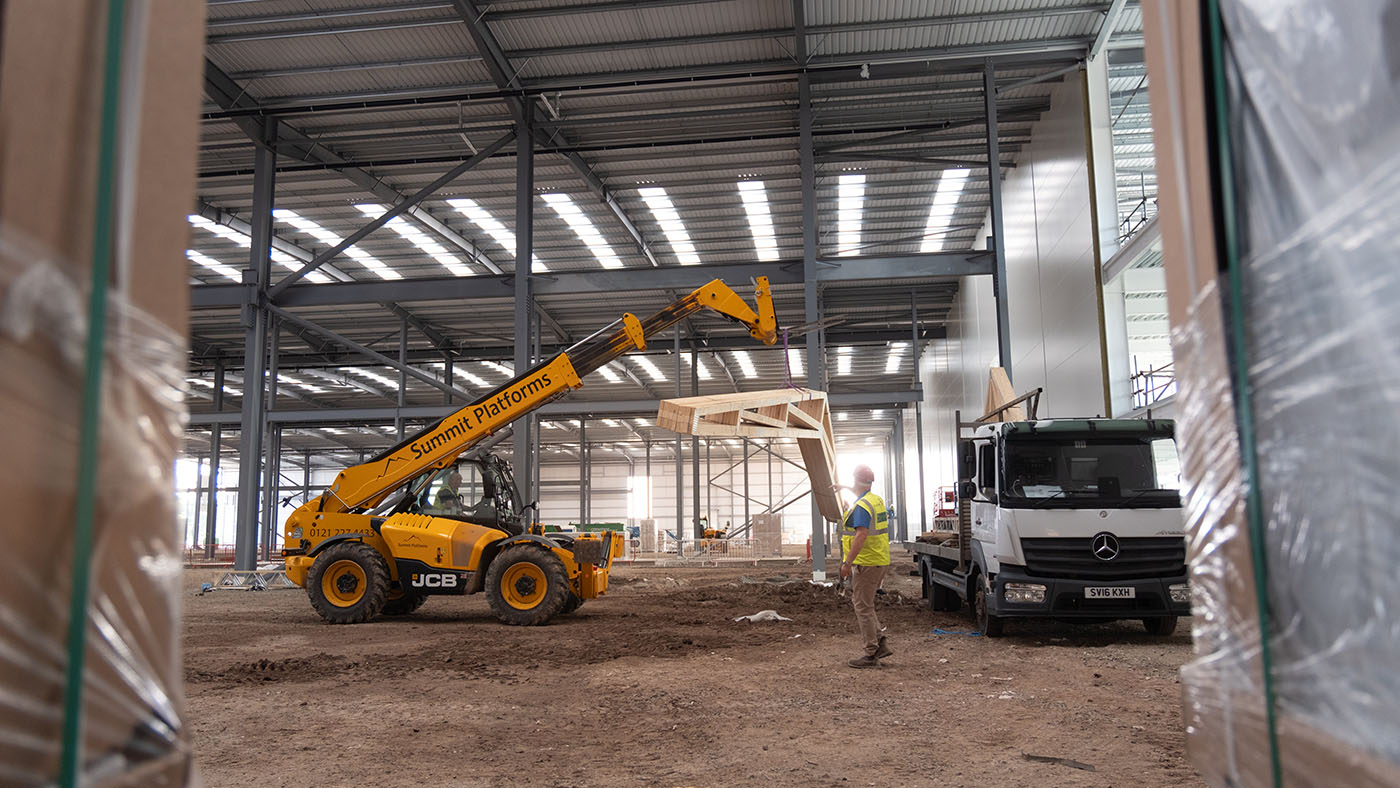 Yellow JCB teleporter lifts a series of scissor trusses off a flatbed lorry inside a factory