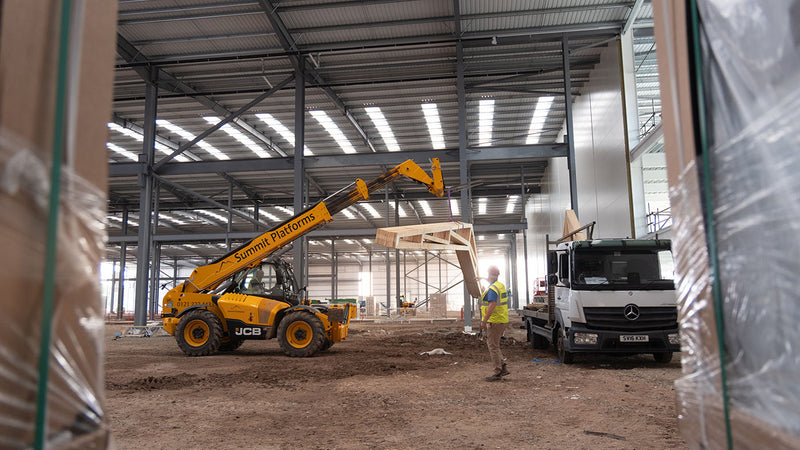Yellow JCB teleporter lifts a series of scissor trusses off a flatbed lorry inside a factory