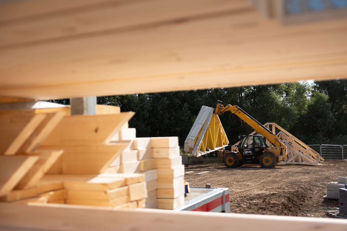 a yellow teleporter moving standard roof trusses across a gravel yard, with timber in the foreground and trees in the background