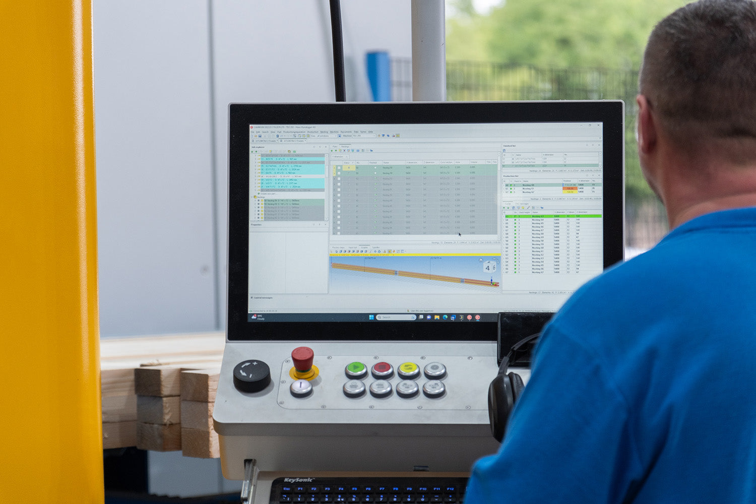 A man in a blue shirt sits at a control console with a computer monitor showing an industrial software interface, with a yellow machine guard visible on the left.